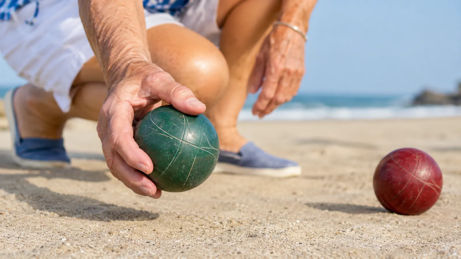 An older woman holding a bocce ball, about to roll it on packed sand