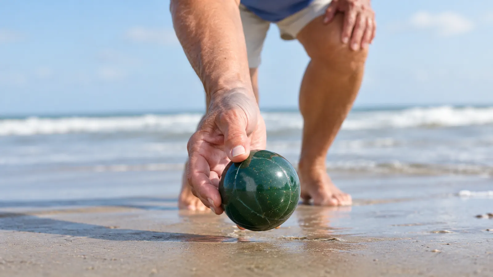 A hand holding a bocce ball while tossing it underhand on a flat stretch of wet sand near the water
