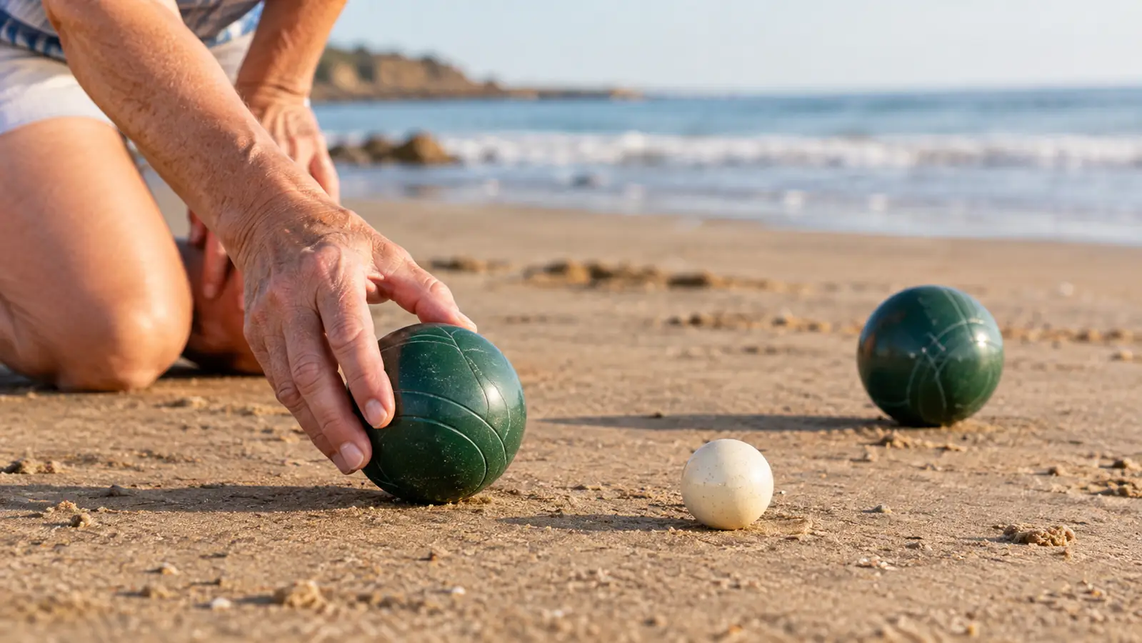 An older woman rolling a bocce ball across packed sand toward the small target ball, with another ball resting nearby