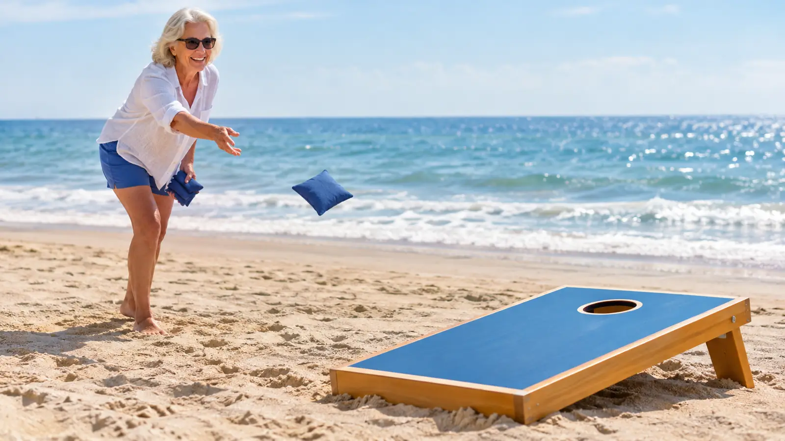 An older woman tossing a bean bag toward a portable cornhole board on the sand