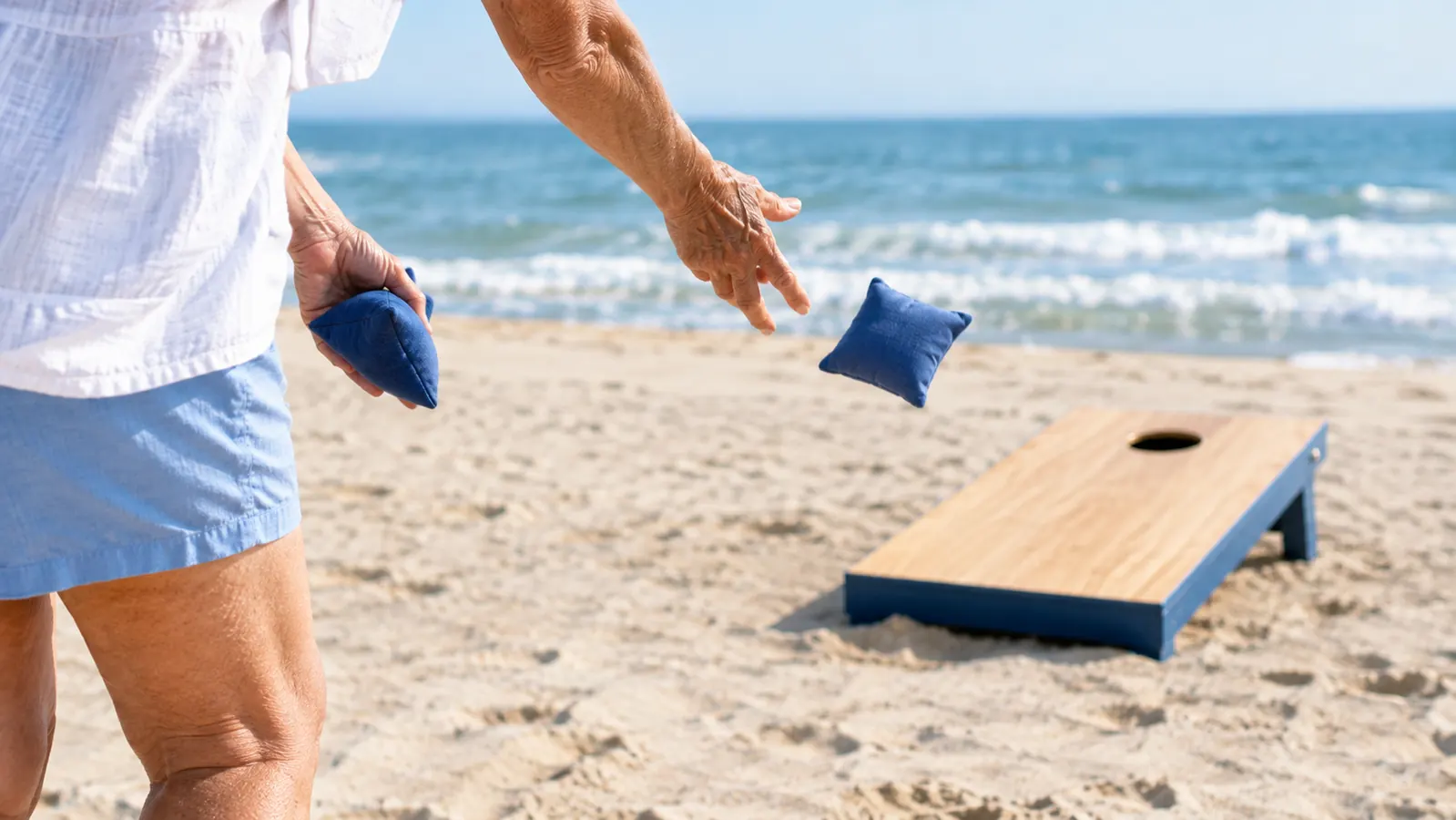 An older woman tossing a bean bag underhand toward a cornhole board set up on beach sand
