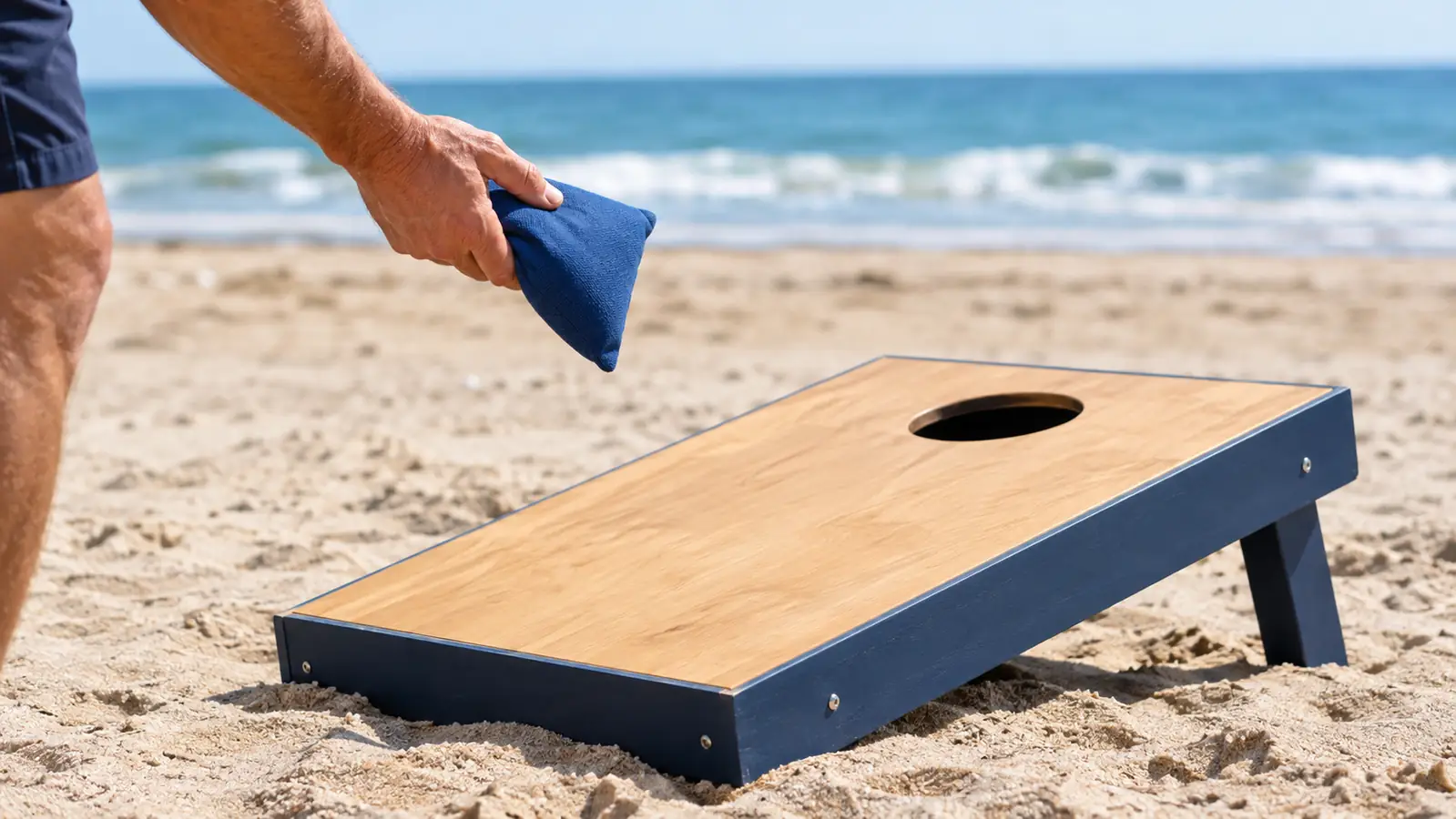 A hand holding a bean bag above a cornhole board sitting on packed beach sand