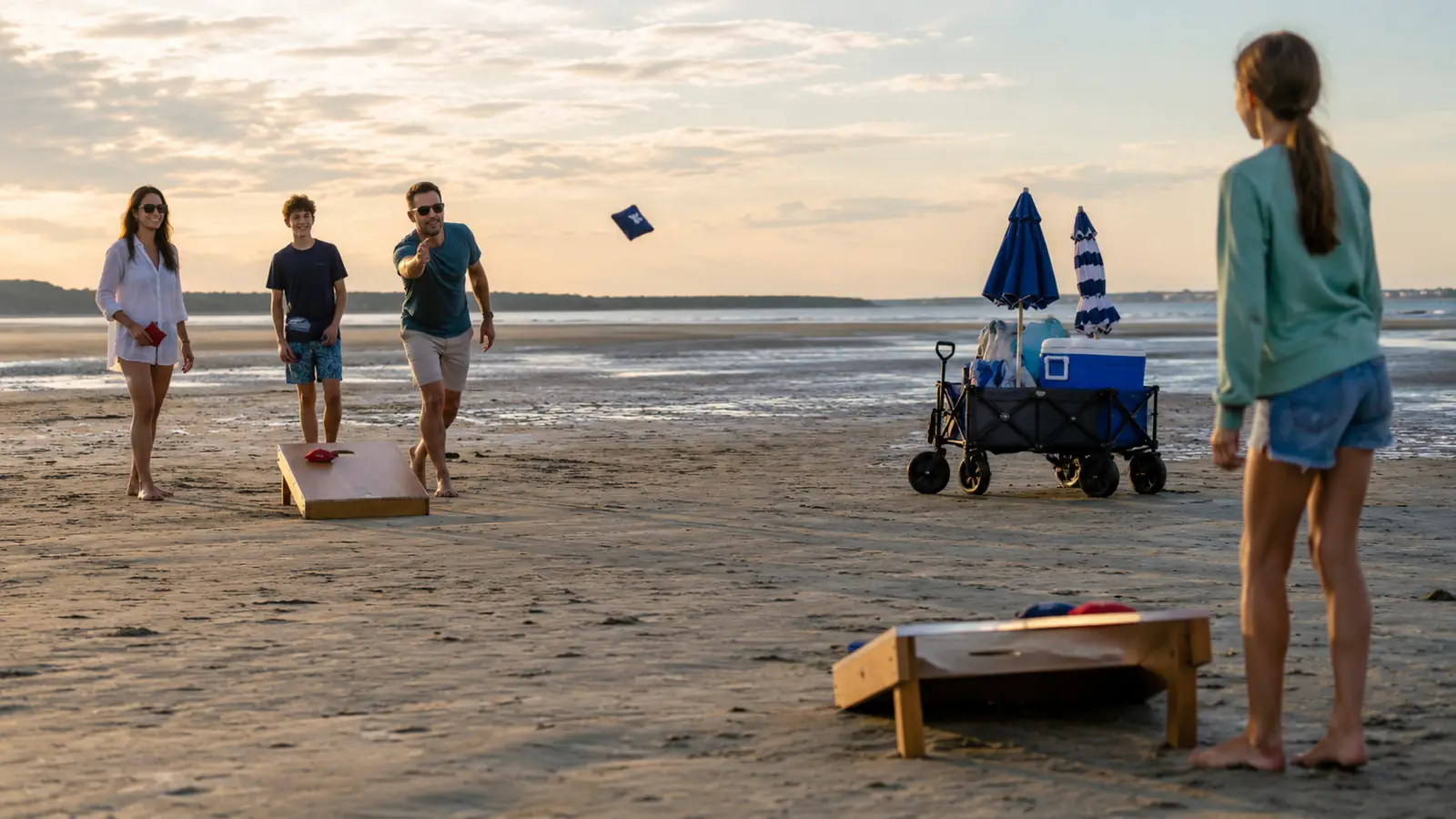 A family playing cornhole on flat sand at low tide with a wagon, umbrellas, and a cooler set up nearby
