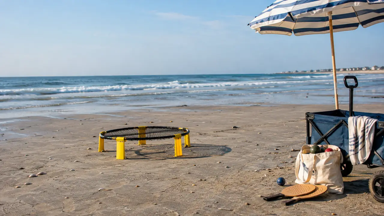 A roundnet set, a wooden paddle set and a bag of bocce balls on flat sand at low tide beside a striped umbrella and a beach wagon