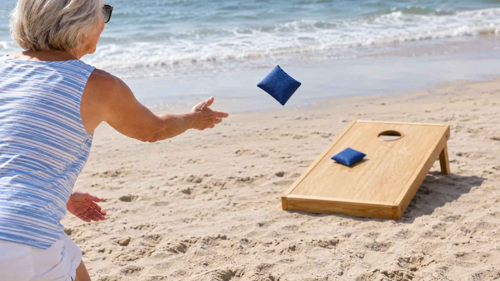 An older woman's hands tossing a bean bag underhand toward a cornhole board set up on flat sand