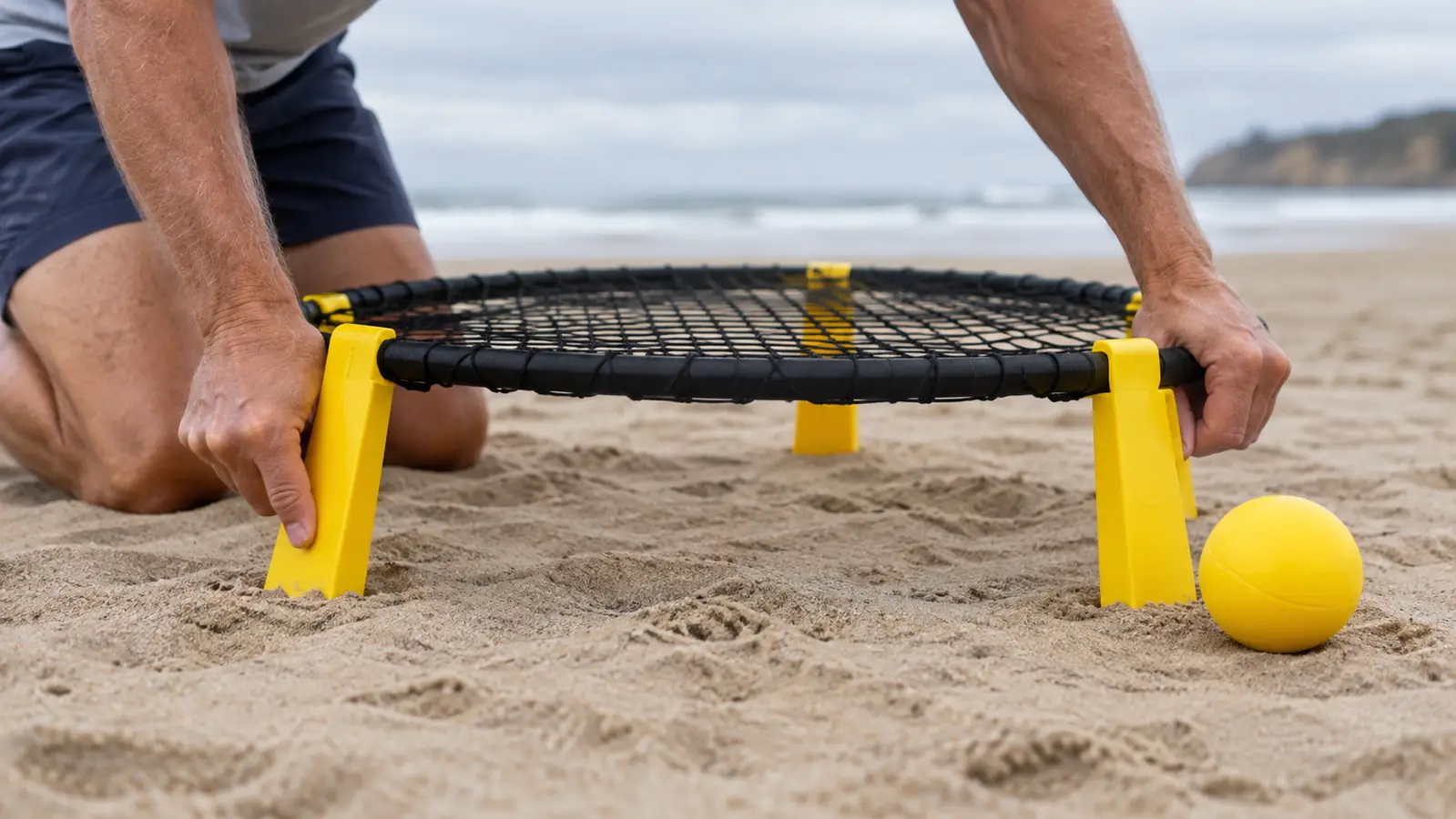 Close view of hands setting a roundnet net and ball into flat sand