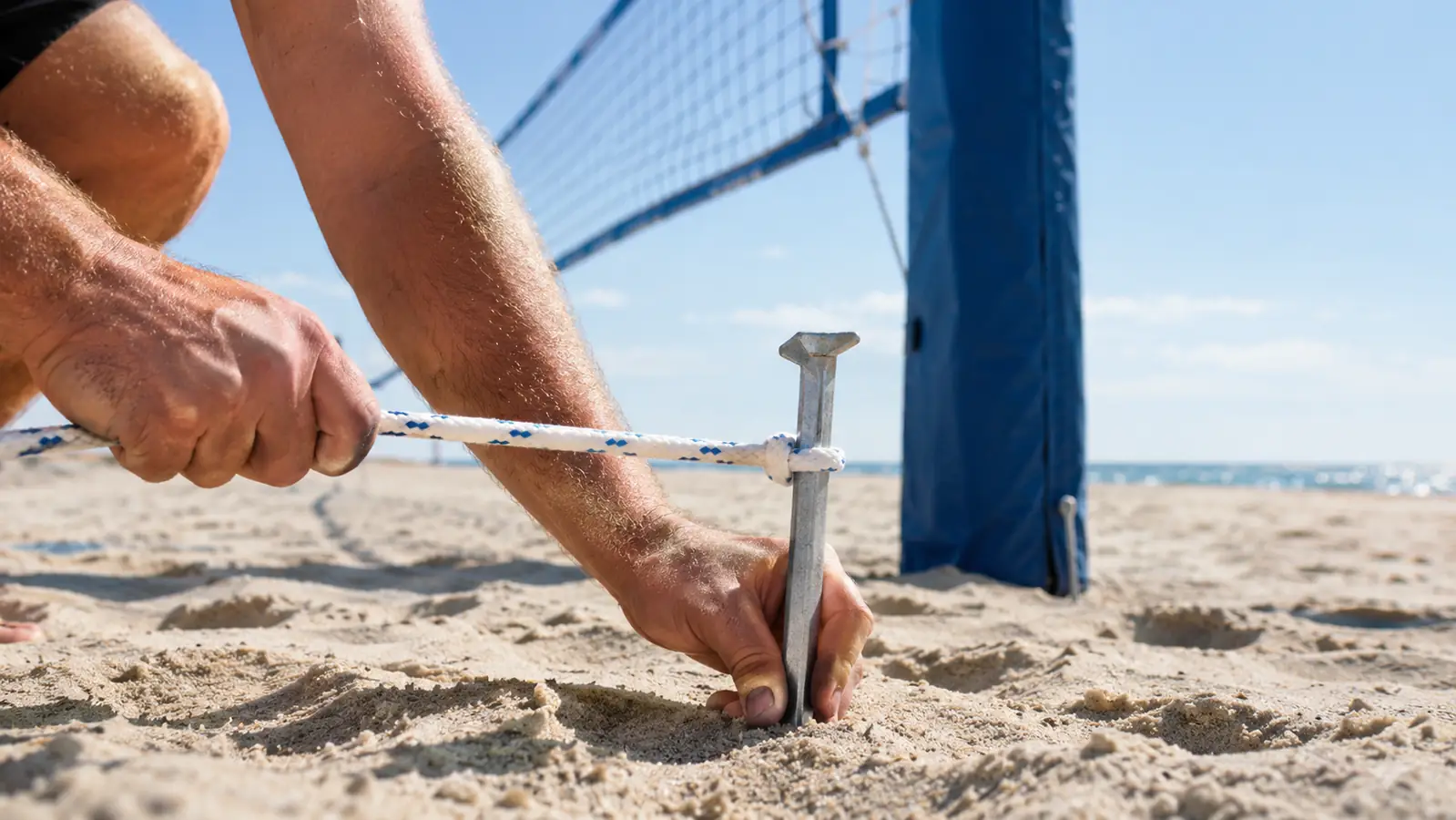 Hands pulling a guide rope taut beside a volleyball net pole anchored in the sand