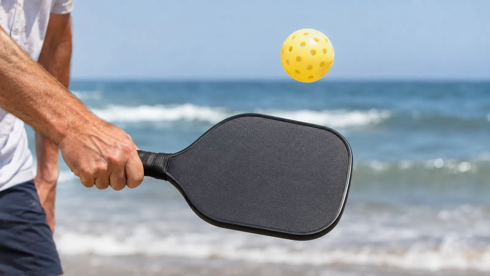 A hand swinging a paddle with the ball in the air above it, the ocean blurred in the background