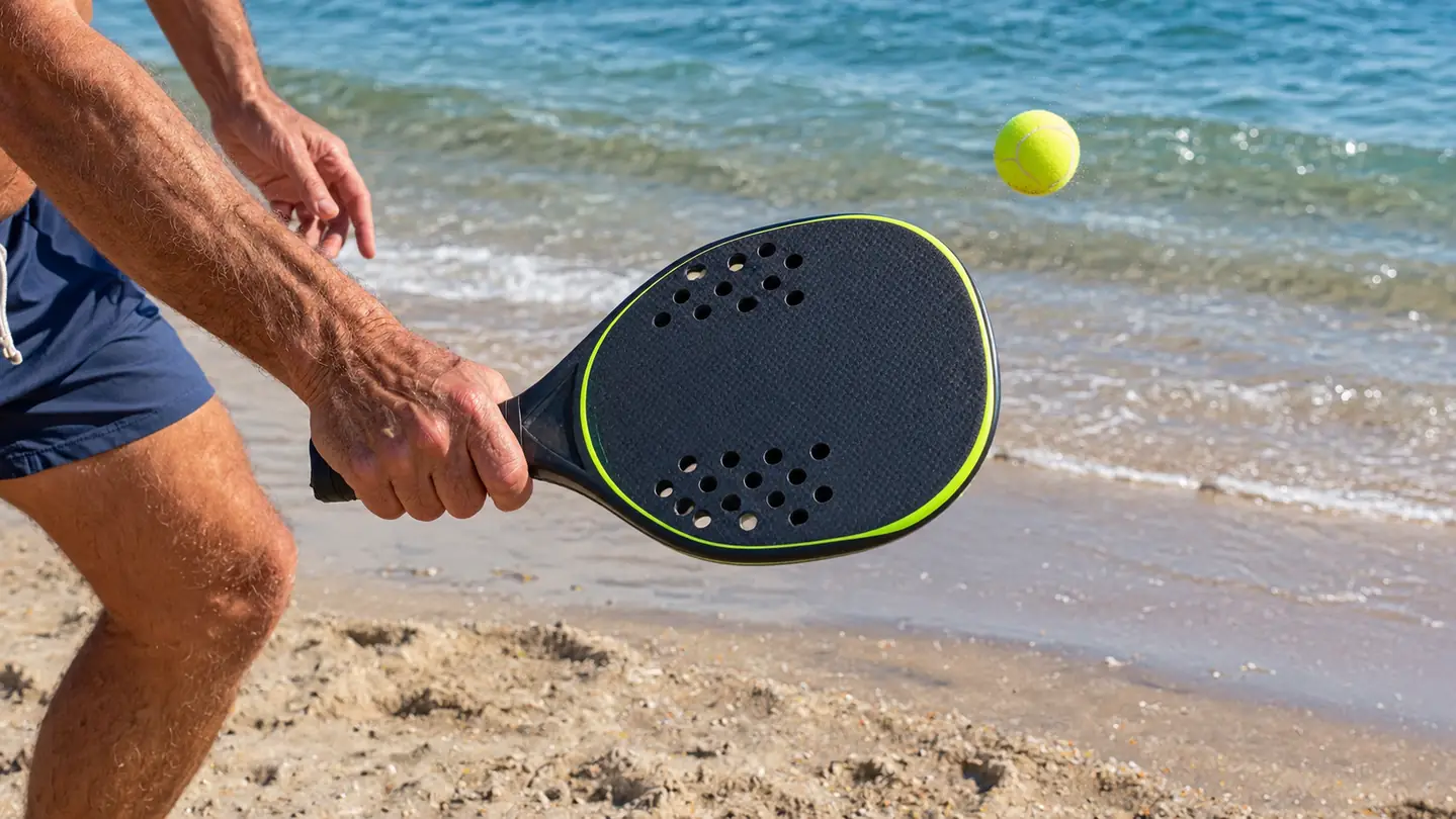 A close view of a hand gripping a paddle mid-swing with the ball in the air above the sand
