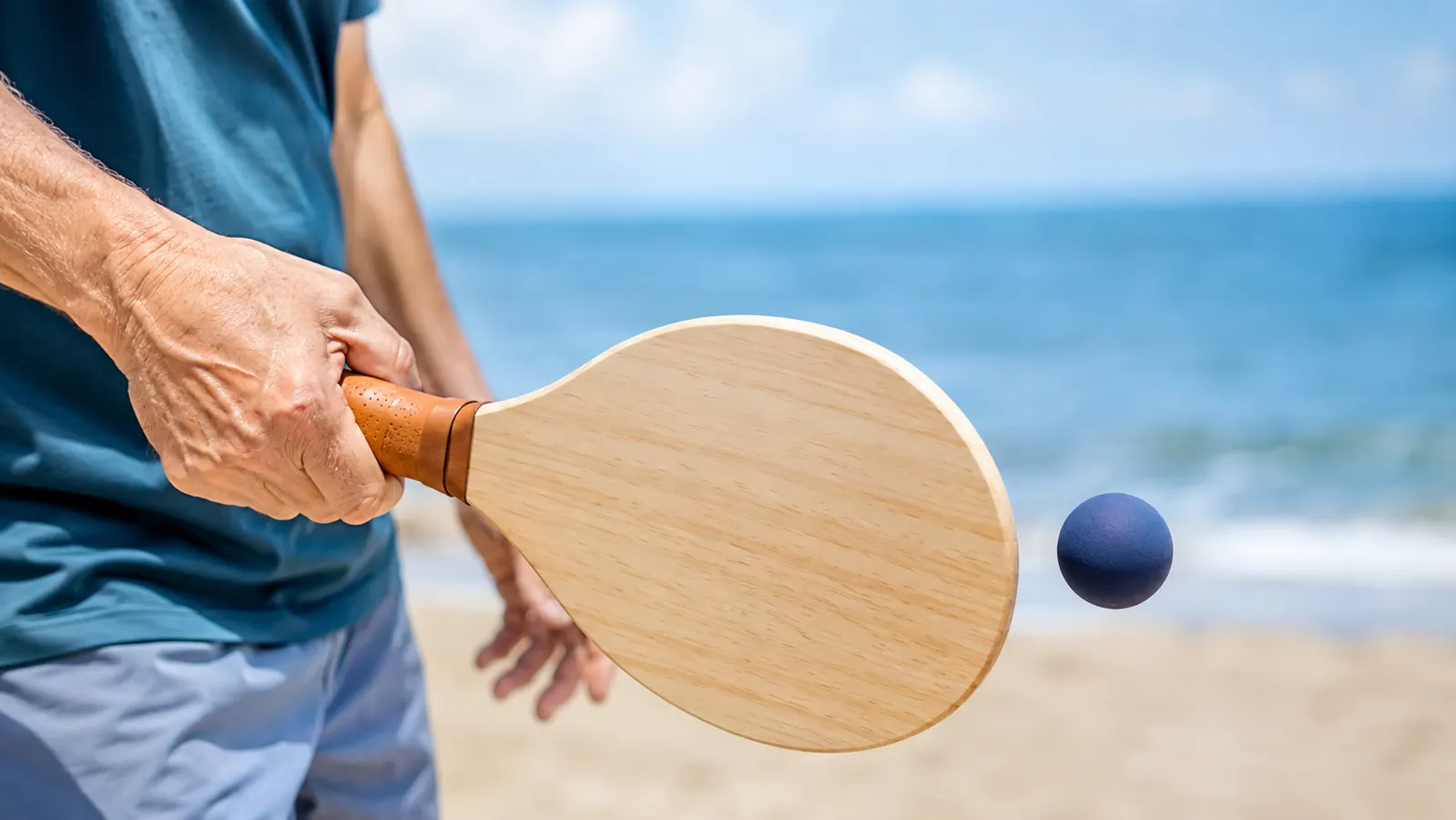 A hand gripping a wooden paddle mid-swing, the small ball just above the paddle face, ocean and sand softly blurred behind
