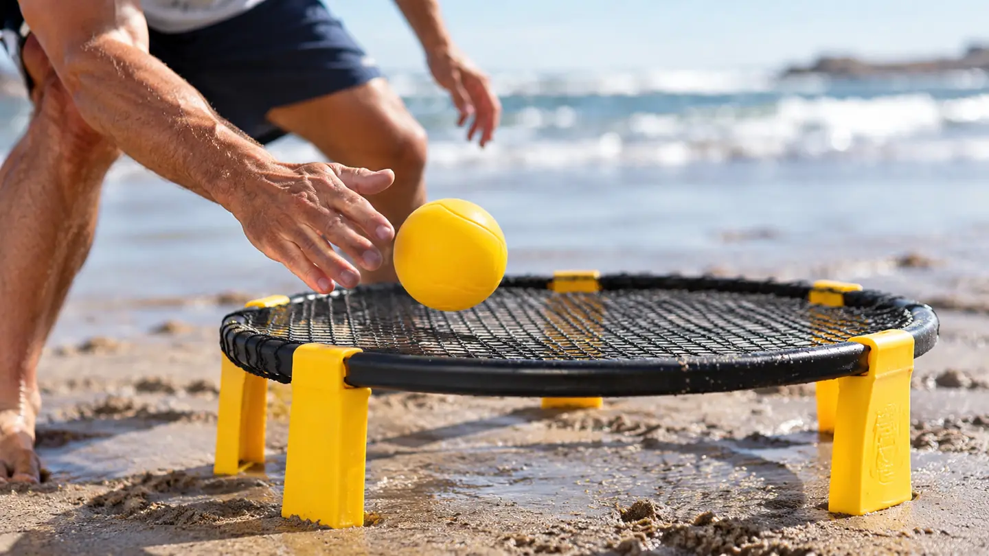 A close view of a player's hand coming down on the ball where it sits on the taut net, the roundnet frame planted on packed sand