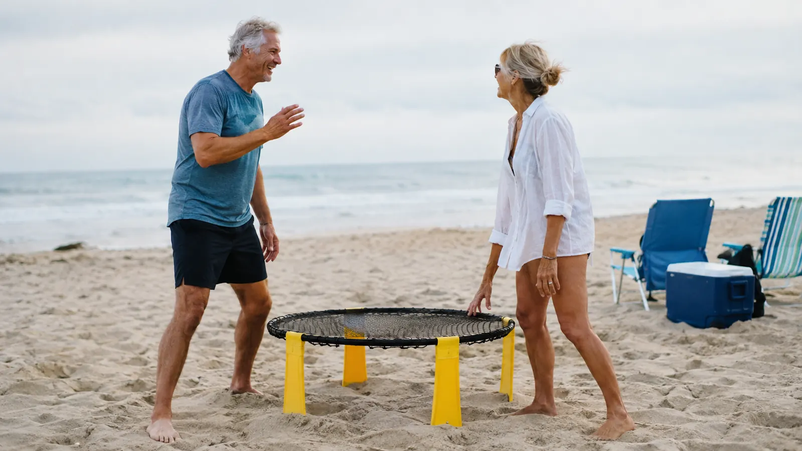 An older couple facing each other across the roundnet on firm sand, a cooler and beach chairs nearby