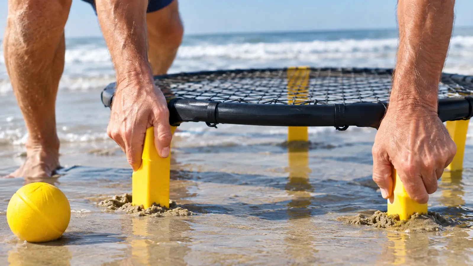 A player's hands pressing a small round net into firm sand near the waterline, with a ball resting on the sand beside it