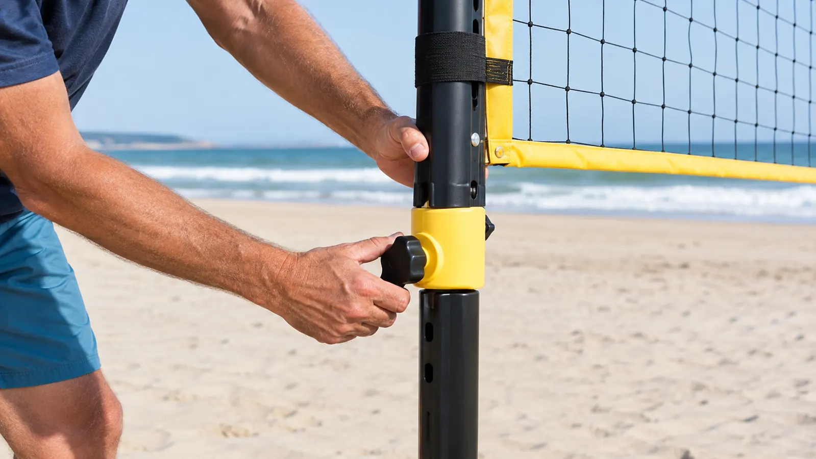 Hands adjusting the height mechanism on a volleyball net pole standing in the sand, net taut, ocean in the background