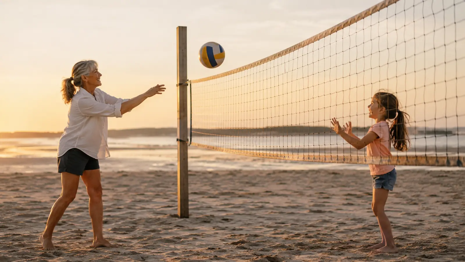 An older woman on one side of a volleyball net tossing a ball to a child on the other side, flat sand and low tide