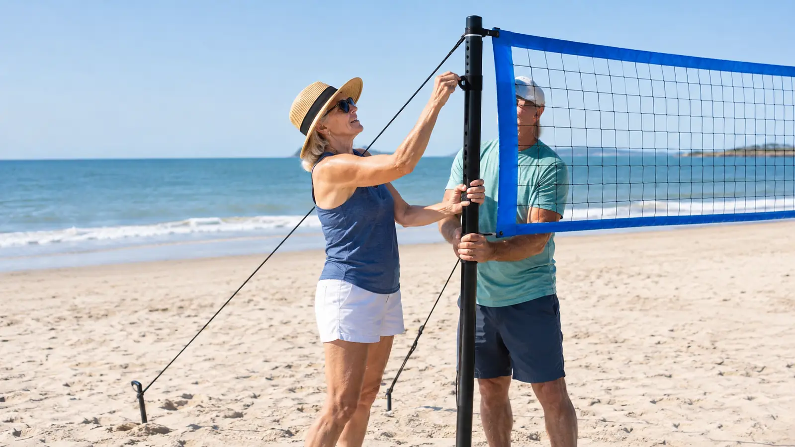 An older woman adjusting the height of a portable volleyball net on the sand while another adult holds the pole steady