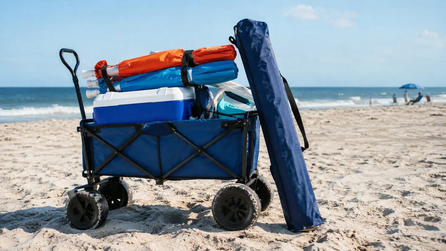 A packed beach wagon holding two umbrellas and a cooler, with a volleyball net carrying bag leaning against it on the sand
